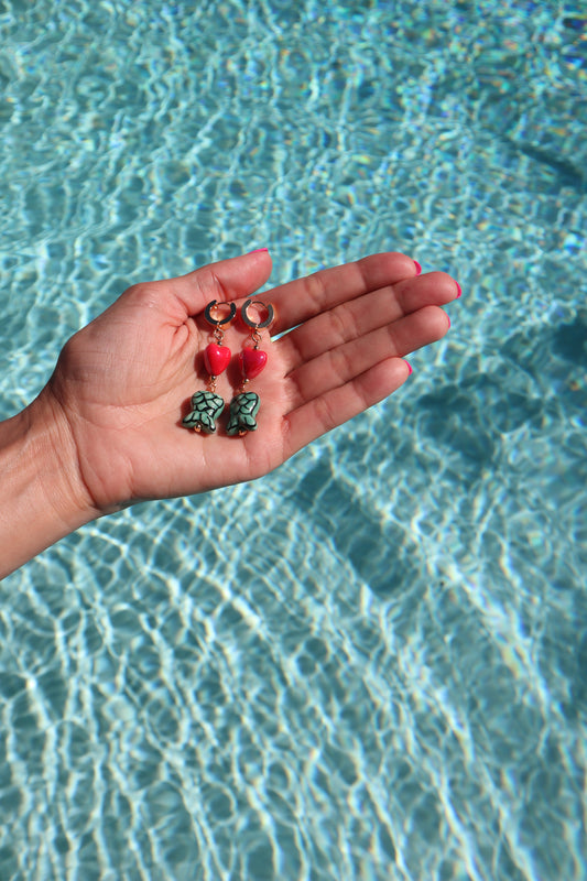 Gold plated huggies with dangling heart and mermaid tail on hand in front of pool