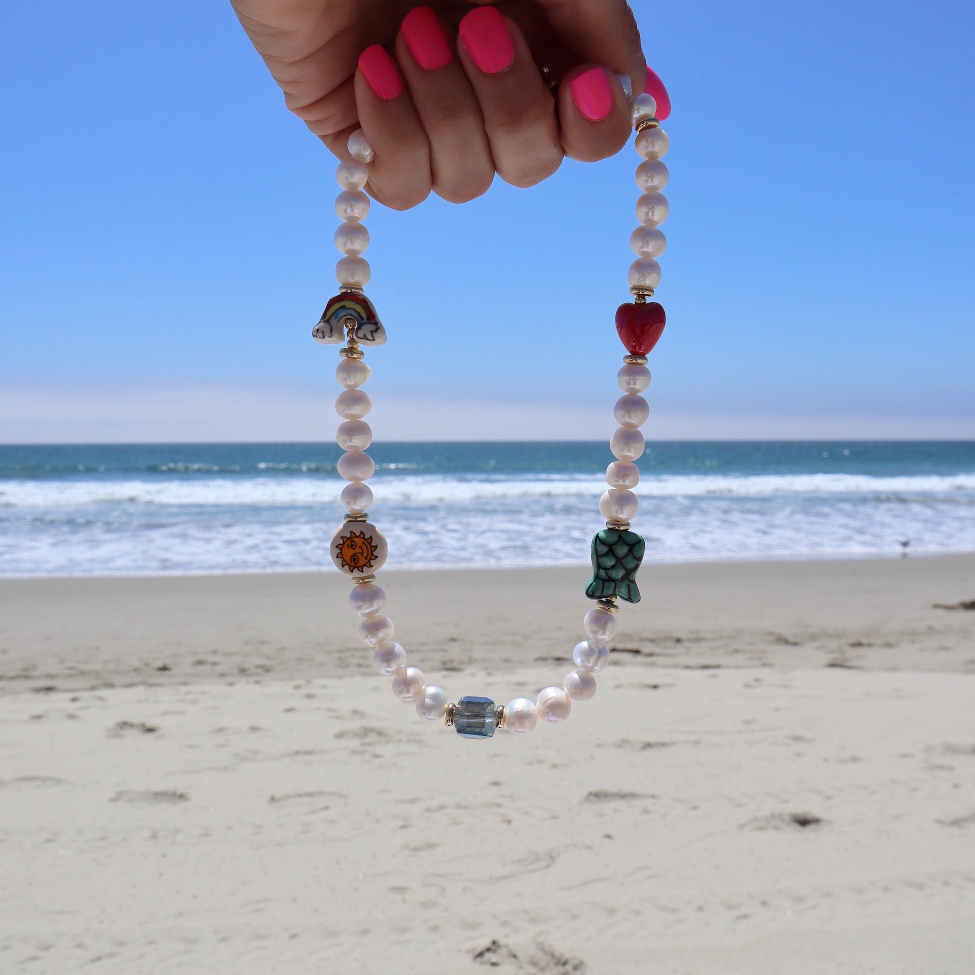 lifestyle-photo-at-the-beach-of-manana-sera-bonito-necklace-hand-made-with-freshwater-pearls-ceramic-heart-mermaid-sun-rainbow-beads-iridescent-blue-glass-stone-by-la-based-latina-artist-nayda-de-jesus