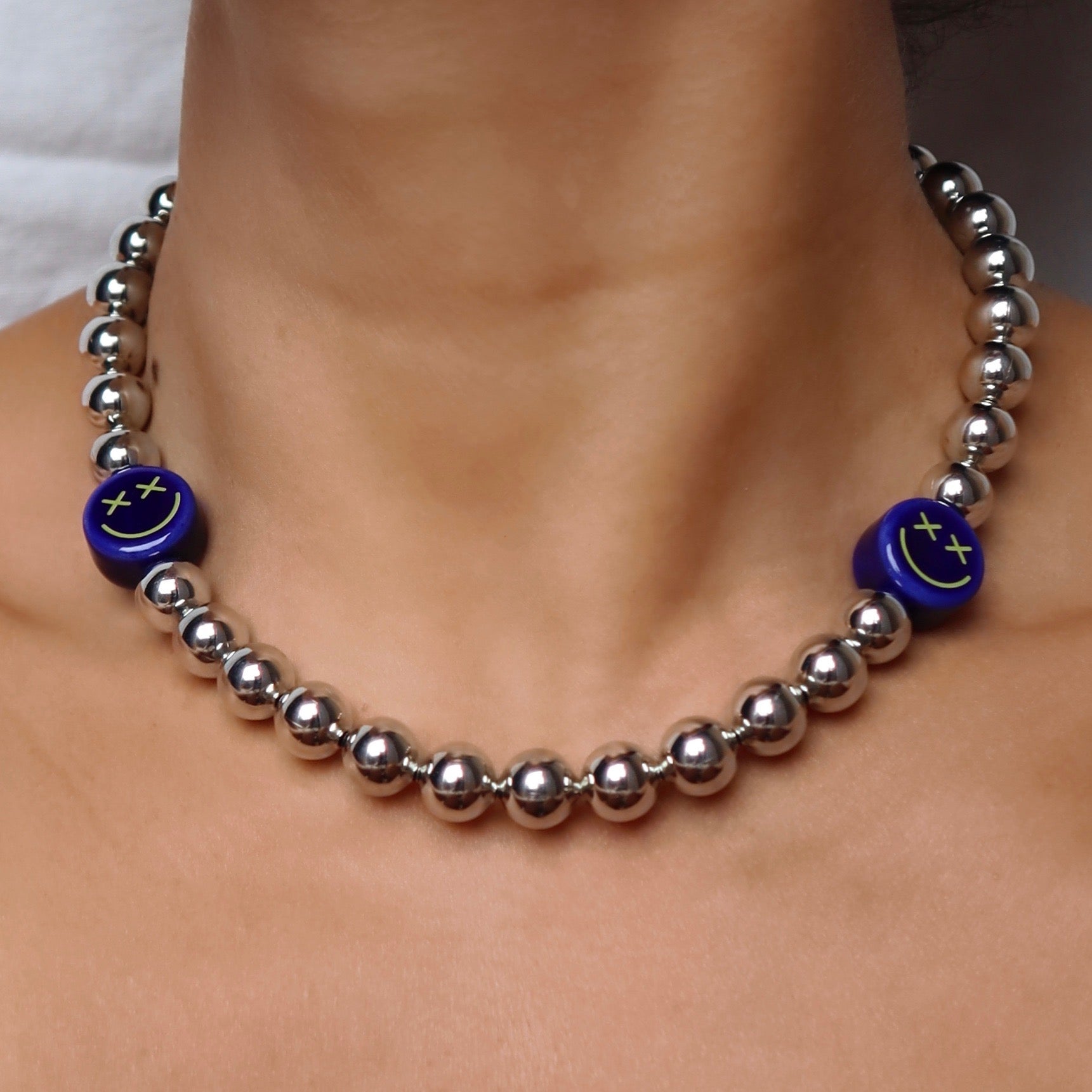 lifestyle-photo-of-en-shock-necklace-made-with-blue-crossed-out-eyes-ceramic-smiley-face-beads-and-silver-metallic-beads-along-18-k-gold-filled-spring-clasp-closure-by-latina-artist-nayda-de-jesus